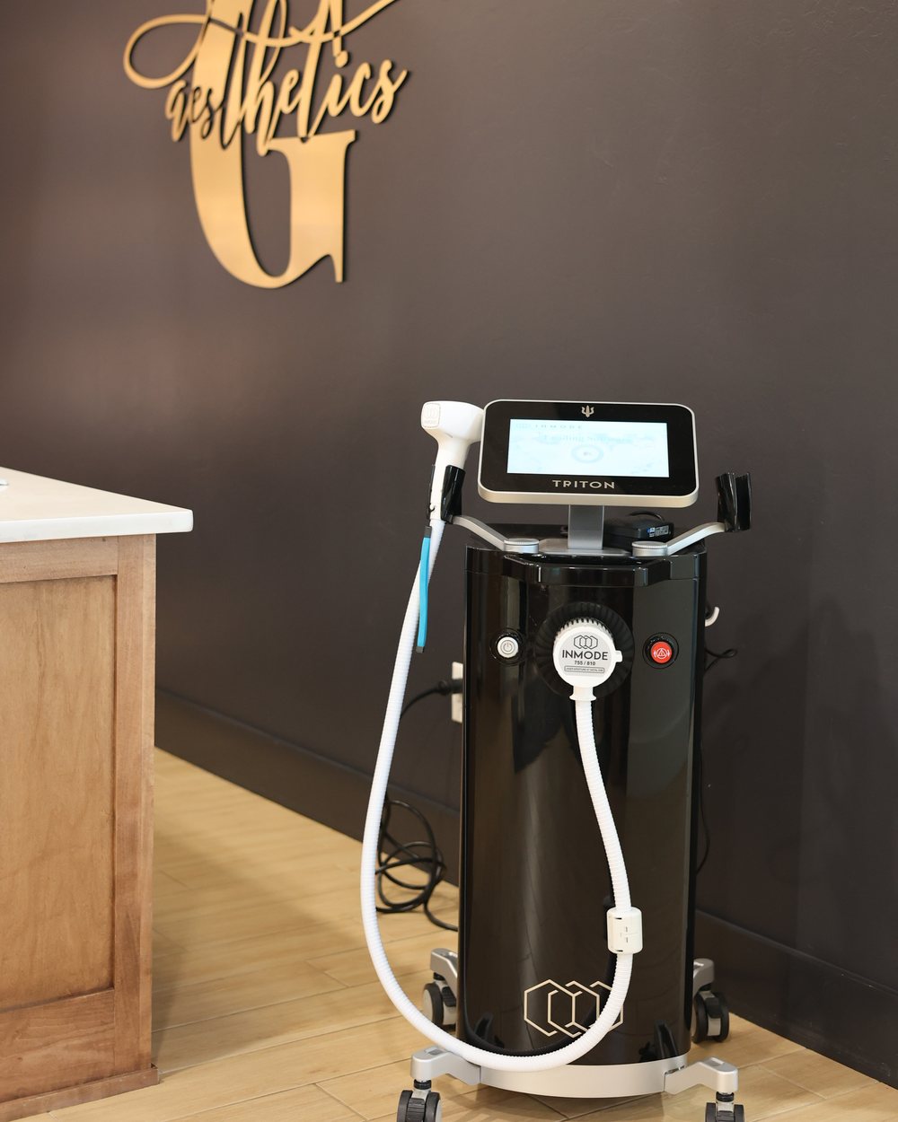 The InMode Triton laser and its handpiece beneath the gold Gudgel Aesthetics wall mark